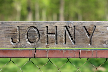 Dilapidated vintage retro sign with carved Johny on top mounted on metal pole of wire fence with green garden in background on warm sunny spring day