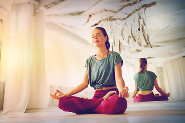 mindfulness, spirituality and healthy lifestyle concept - woman meditating in lotus pose at yoga studio with soft sunlight