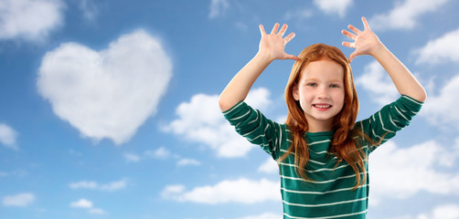 childhood, valentine's day and people concept - smiling red haired girl in green striped shirt having fun and making big ears over blue sky and heart shaped cloud clouds background