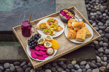 Breakfast on a tray with fruit, buns, avocado sandwiches, smoothie bowl by the pool. Summer healthy diet, vegan breakfast. Tasty vacation concept