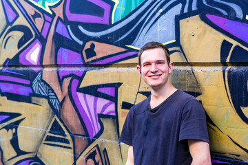 Jugendlicher Mann lächelt freundlich mit Kopfhörern im Ohr an einer Graffiti Mauer stehend