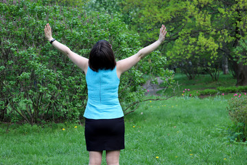 Woman in blue blouse and black skirt stands with her arms outstretched in a summer park, back view. Harmony with nature, concept of romantic vacation, ecology, dreaming, joy, positive emotions