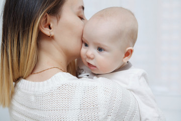 Young happy loving mother plays kisses her baby daughter in her arms against the window. Maternal care. Childcare. Close-up.