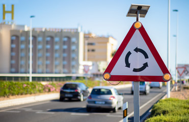 A triangular warning sign with flashing lights stands in front of a roundabout on a sunny day in Spain and cars drive towards the circle.