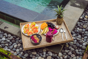 Breakfast on a tray with fruit, buns, avocado sandwiches, smoothie bowl by the pool. Summer healthy diet, vegan breakfast. Tasty vacation concept