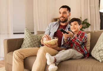 family, childhood, fatherhood, technology and people concept - happy father and little son with popcorn and remote control watching tv at home in evening