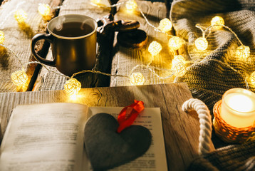 Holidays mood photo. Christmas lights and warm knitted sweater. Hot tea mug with a book for cosy evening. Sweet gingerbread and wooden heart on tray. Perfect winter flat lay with candle. Hygge concept