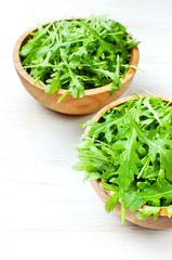 Fresh green arugula leaves on wooden bowl, rucola salad on white wooden rustic background with place for text. Selective focus. Rocket salad or arugula, healthy food, diet. Nutrition concept.