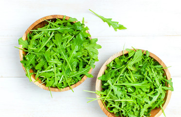 Fresh green arugula leaves on wooden bowl, rucola salad on white wooden rustic background top view with place for text. Rocket salad or arugula, healthy food, diet. Nutrition concept.