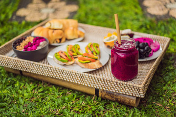 Breakfast on a tray with fruit, buns, avocado sandwiches, smoothie bowl standing on the grass