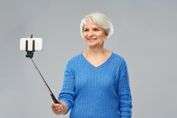 technology and old people concept - smiling senior woman taking picture by smartphone on selfie stick over grey background