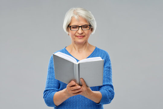 vision, wisdom and old people concept - smiling senior woman in glasses reading book over grey background