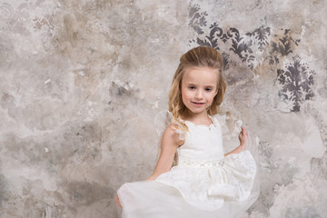 Portrait of a little attractive smiling girl in a white dress with curled hair against the background of a grunge wall.