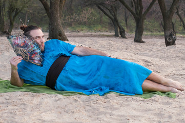 handsome tricky man with hair bun  and blue kimono lying with fun and looking at camera on sand on beach