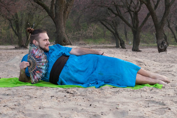 transsexual handsome man with make up, hair bun lying on sand in blue kimono, holding hand fan and...