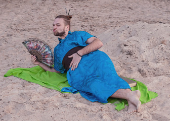 transsexual handsome man with make up, hair bun lying on sand in blue kimono, giving kiss, holding...