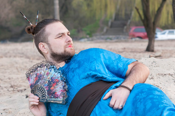 handsome tricky bearded man with hair bun and blue kimono lying with fun and looking away on sand...