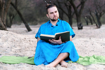 Obraz premium serious handsome bearded man in blue kimono sitting, holding opened large book and looking away on sandy beach near trees at spring