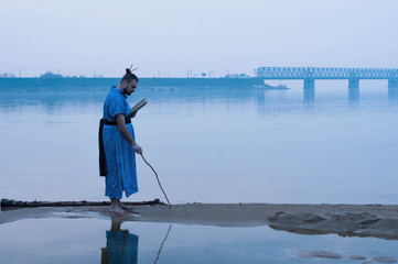 Naklejka premium side view of overweight bearded man in blue kimono standing on river bank and writing on sand with wooden stick and holding book in front of bridge