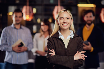 Portrait of beautiful Caucasian blonde businesswoman standing at restaurant with arms crossed. In background her colleagues standing and posing.