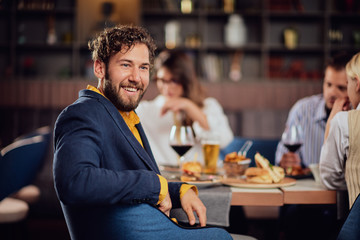 Young smiling man dressed smart casual sitting at restaurant and looking over shoulder. In background his friends having dinner.