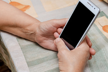 Hand old woman holding white smartphone with black screen on the background of the table at home. A modern retired elderly woman uses a mobile phone, earning money through the Internet