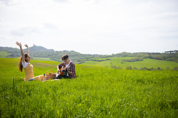 A young couple in bright clothes sitting on a bright green meadow and the man playing guitar while his girlfriend having fun