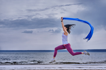 Young slim brunette woman jumping while jogging while holding a blue scarf in her hands. A woman is engaged in gymnastics in the spring morning on the sandy bank of a large river. Cloudy morning.