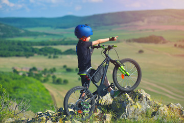 Fototapeta na wymiar Little child stand next to his mountain bike on mountains edge and looks at the beautiful scenery