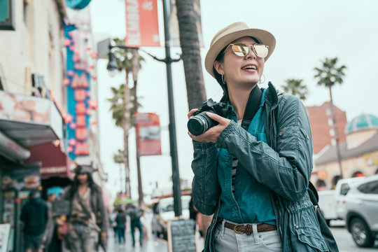 young asian female photographer holding slr camera smiling taking picture on street outdoor sunny day. happy woman traveler in sunglasses walking on hollywood boulevard take photo for memory trip usa