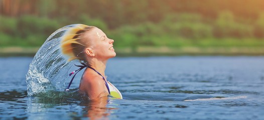 Active young blonde woman waving hair splashing water in river. Beautiful healthy lady relax and laughing, raising head out of the water. Vacation in paradise enjoying swimming. Motion freeze.