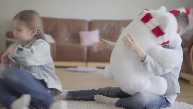 Portrait twin boy and girl sitting on the floor in living room and dont know how to share between them a big taddy bear. Kids pulling toy to each other to own it