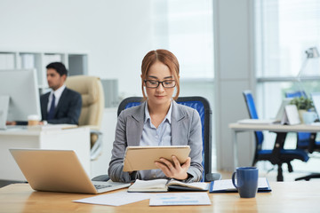 Young businesswoman in eyeglasses sitting at her workplace and concentrating on her work she using digital tablet for online work with businessman working in the background