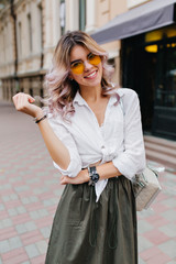 Amazing young woman in long skirt and classic white shirt enjoying free time outside and posing with pleasure. Outdoor portrait of stylish curly lady in elegant wristwatch walking by cafe.
