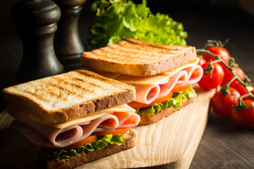 Close-up of two sandwiches with bacon, salami, prosciutto and fresh vegetables on rustic wooden cutting board. Club sandwich concept.