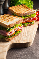 Close-up of two sandwiches with bacon, salami, prosciutto and fresh vegetables on rustic wooden cutting board. Club sandwich concept.