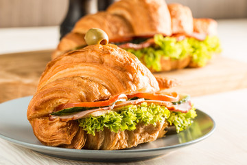 Photo of tasty and fresh croissant sandwich with salad, ham, cheese, tomatoes on wooden background. Morning breakfast concept. Healthy and fast food.