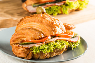 Photo of tasty and fresh croissant sandwich with salad, ham, cheese, tomatoes on wooden background. Morning breakfast concept. Healthy and fast food.
