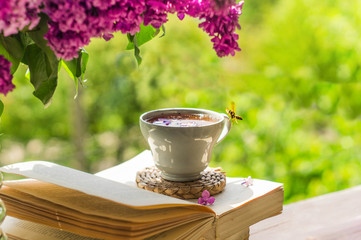 Book, glasses, cup of tea and lilac on a wooden window. The bee flies beautifully over the lilac...