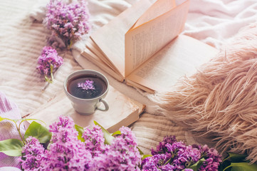 Still life details in home interior of living room. Sweaters and cup of tea with lilac flowers and spring decor on the books. Read, Rest. Cozy spring concept.