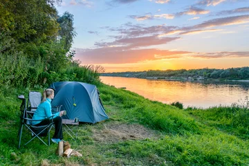 Fotobehang Jonge vrouw geniet van de zonsondergang in de reisstoel naast de tent op de camping bij de rivier © Maks_Ershov