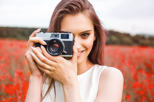 cheerful young girl in a summer dress is holding an analog film camera in front of a colorful, blooming field, concept of creativity and self-expression