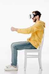 side view of young man in vr headset imitating driving while sitting on chair on white background