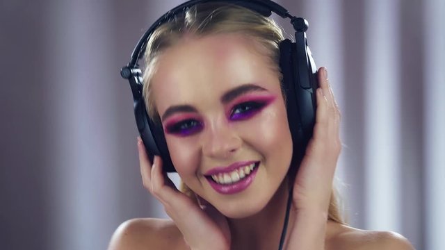 Fashion model woman with trendy pink make-up listening music in headphones. Glitter vivid makeup. Studio video