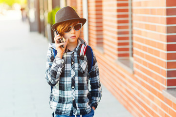 Stylish teenage boy with mobile phone. Hipster kid in sunglasses and modern hat at city street. Young boy talking on the smartphone outdoors.