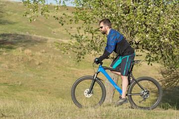 Obraz premium Cyclist in shorts and jersey on a modern carbon hardtail bike with an air suspension fork rides off-road on green hills near the forest 