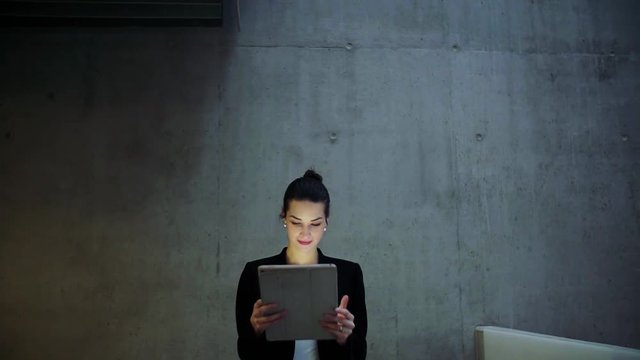 Young business woman with tablet walking in office, concrete wall in background.