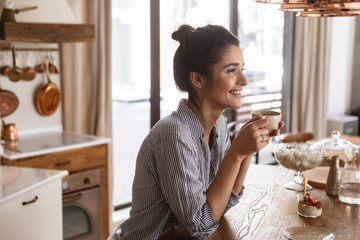 Photo of happy brunette woman 20s drinking coffee while having breakfast at home - obrazy, fototapety, plakaty