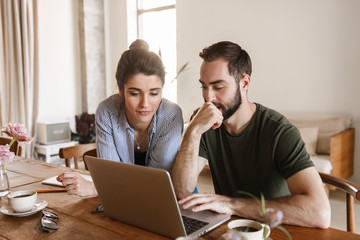 Image of concentrated brunette couple working on laptop together while sitting at table at home