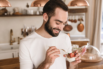 Portrait of positive brunette man 20s eating panna cotta dessert while having breakfast in kitchen...